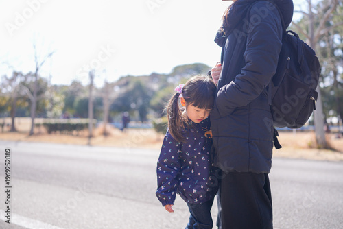 Sunlit street beside a quiet park. Mother hugs child warmly beside road. Little girl looks down, shy and quiet. Both wear dark coats, matching winter style. Soft light casts gentle glow on their forms