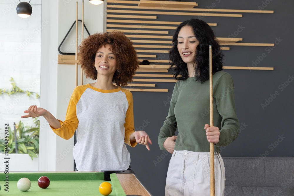 Fototapeta premium Two female friends playing pool in raglan and olive tops, smiling and holding cue with balls