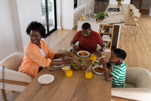 African American family with child sitting at wood table in modern kitchen, sharing pasta bowls
