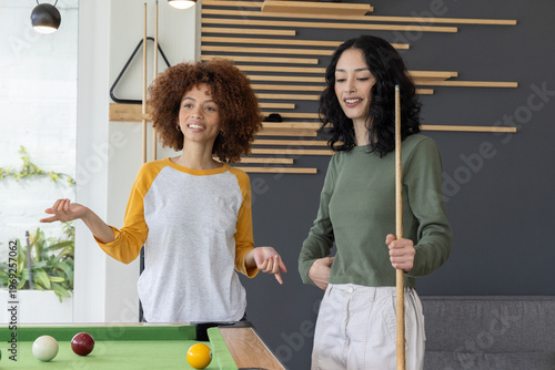 Two female friends playing pool in raglan and olive tops, smiling and holding cue with balls