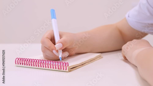 Person is writing in a spiral notebook. The pen is blue and white. The notebook is pink