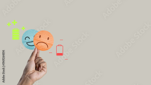 Conceptual image of emotional health featuring a hand holding a sad emoji over a happy one. Icons of full and empty batteries illustrate energy levels and mental exhaustion
