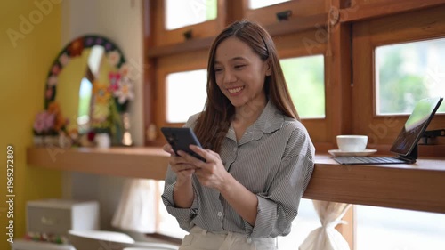 A woman is looking at her cell phone while standing in front of a window. She is focused on her phone and not paying attention to her surroundings
