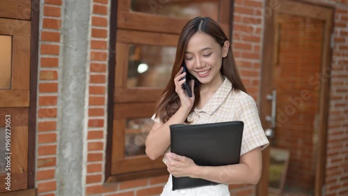 A woman is talking on her cell phone while holding a black folder. She is smiling and she is in a good mood