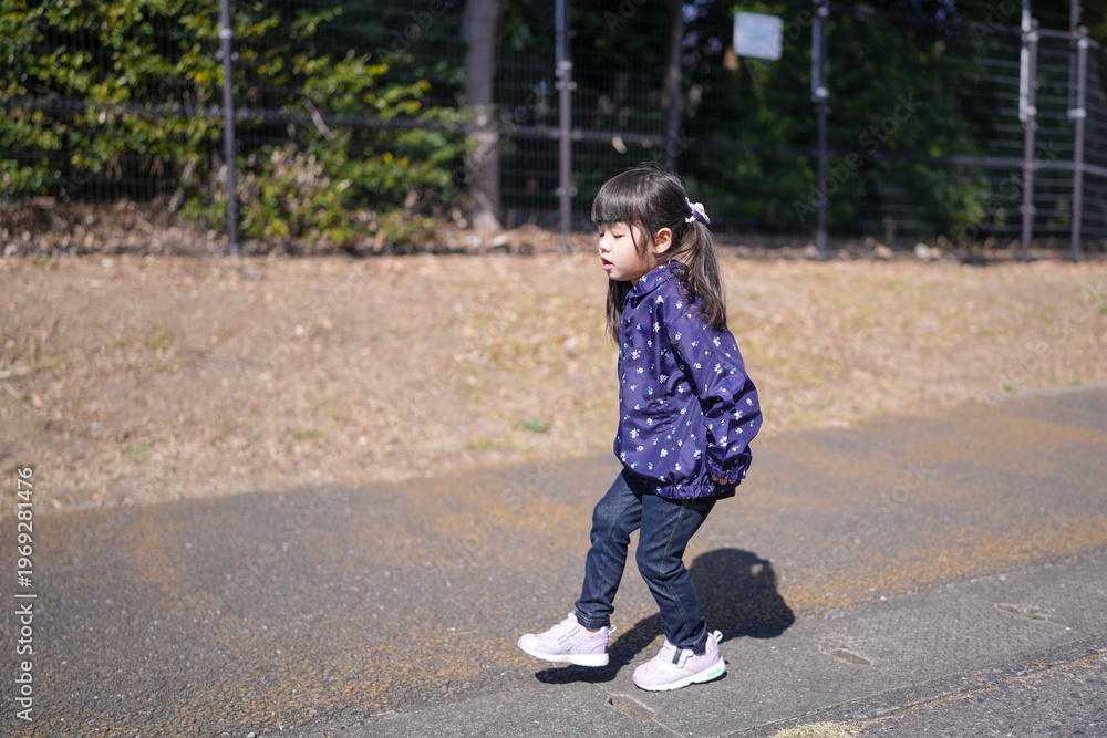 Fototapeta premium Little girl walks calmly on paved path. Purple jacket adds cozy contrast to bright day. Her hair tied back, she moves with quiet confidence. Background shows dry grass and fence, sunlit outdoors