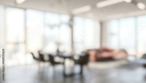 A blurred interior view of a modern office space featuring large windows, comfortable seating, and a table, conveying a professional atmosphere.