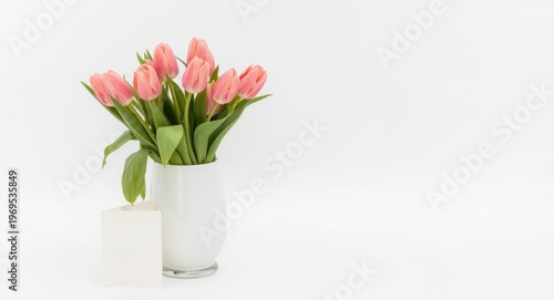 White glass vase filled with blooming pink tulips paired with a greeting card offering copy space on white backdrop