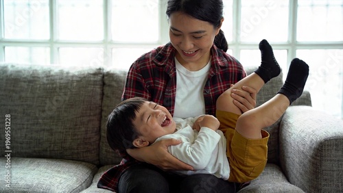 Happy Asian mother playing with her laughing baby son on the sofa at home family bonding and parenting concept joyful child playing on mother lap motherhood love care indoor