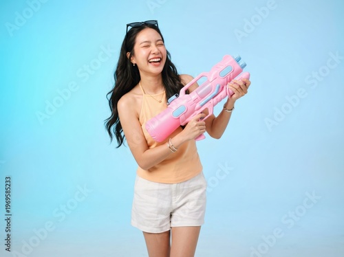 Young asian woman holding pink water gun laughing on blue background wearing summer clothes and sunglasses on head. Studio portrait with copy space. Songkran water festival and summer vacation concept