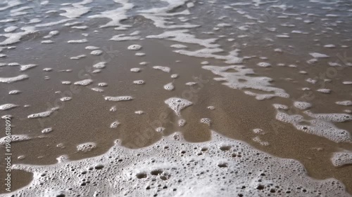 Ocean waves gently wash over sandy beach, creating foam patterns and ripples on the shore, capturing the serene beauty of coastal nature in bright sunlight