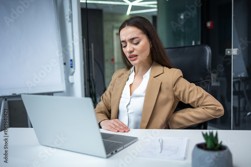 Woman with hip, back, spine spasm, cramp and pain, working from office troubles and issues.incorrect posture concept. frowning woman sitting at desk in front of laptop