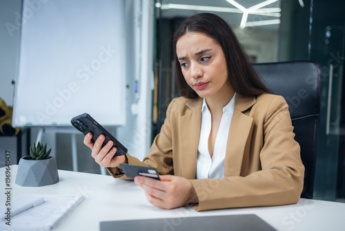 Fraud. Upset young beautiful woman holding a credit card. A disappointed young lady lost money. Sitting at the desk in the office