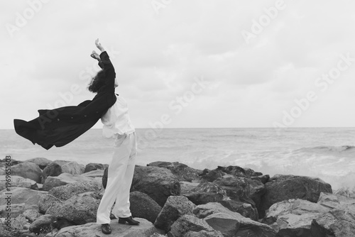 Two figures on rugged rocks by the sea raise arms and fabric in motion, wind lifting garment, black and white image capturing freedom, balance, and connection with nature in a dramatic seaside moment