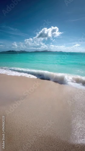 Clear turquoise ocean water washes onto a golden sandy beach under a vibrant blue sky dotted with fluffy white clouds. Sun glistens on the ripples, revealing the shallow seabed.