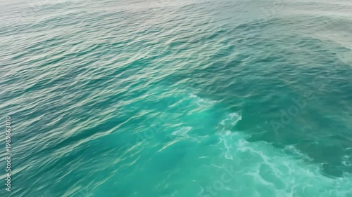 Ocean waves gently ripple across the surface, revealing varying shades of blue and green water, with sunlight reflecting off the tranquil sea in a coastal environment