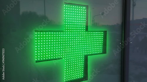 Green illuminated pharmacy cross sign displayed on a glass window, showcasing a bright LED design with a clear urban background during twilight hours