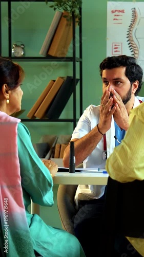 Professional Indian young male doctor consulting with Asian senior retired patients across a table in a modern clinic; healthcare, geriatric assistance, and old age medical support concept.