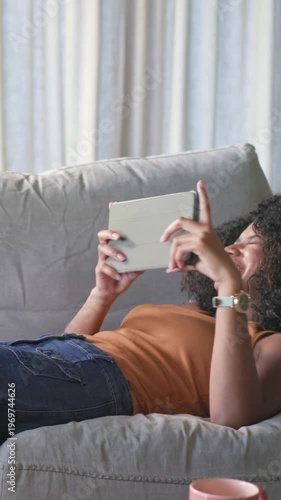 Vertical video: Lying woman in orange top tapping tablet on couch reacting to content near pink mug