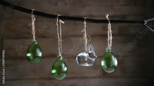Glass Easter decorations transparent bunny and three ribbed green eggs are suspended on rustic-style twine from dark branch against a warm wooden background, creating atmosphere of cozy spring decor