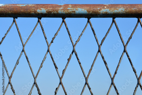 The rusty blue metal pipe and grey wire mesh of a fence show signs of age and corrosion against a clear blue sky