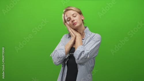 Woman with hands pressed to cheek in sleeping gesture wearing striped shirt on green studio set; serenity.