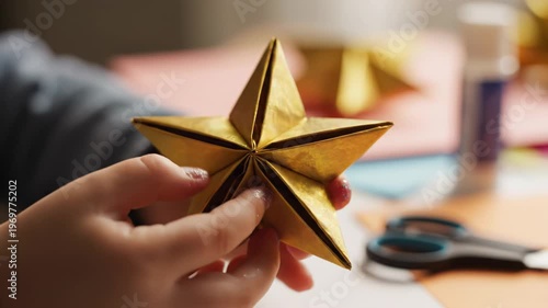 Child's hands carefully assemble a golden paper star ornament, showcasing intricate folds and craftsmanship against a colorful crafting workspace with scissors and glue