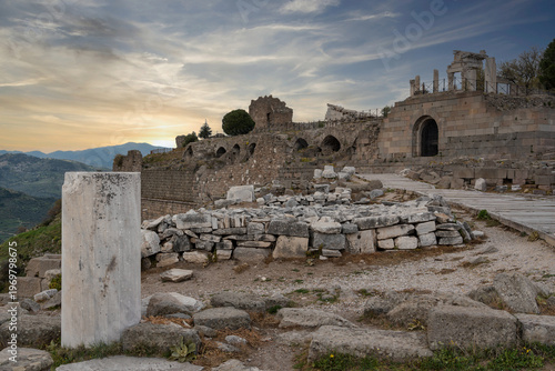 Pergamon Acropolis ruins at sunset, Bergama, Turkey. Ancient stone walls, arches, and column fragments under a dramatic sky, overlooking a mountainous landscape
