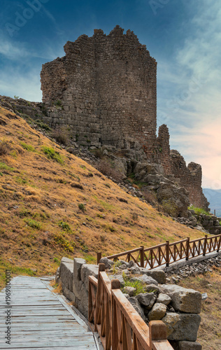 Ancient stone ruins of the Acropolis of Pergamon on a dry grassy hillside with a wooden walkway, Bergama, Turkey under a cloudy blue sky