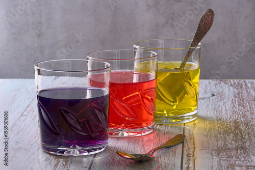 Three glasses filled with different colored jellies are placed on a wooden table. A silver spoon lies beside the glasses