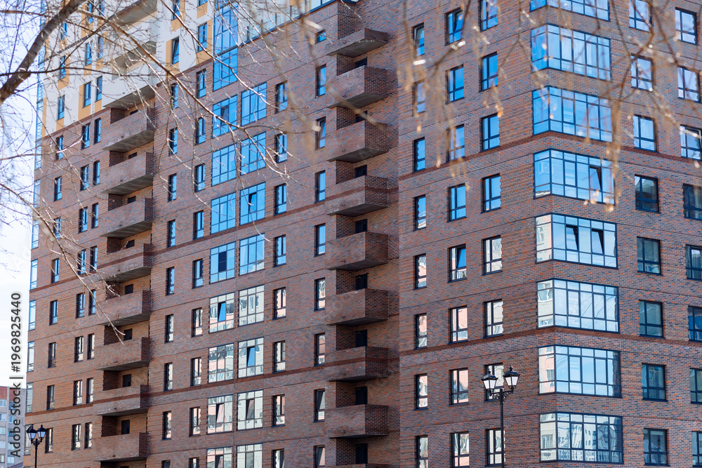 Fototapeta premium Modern apartment building with brick facade and glass windows. Balconies line each floor in repeating geometric pattern. Reflections of sky and trees shimmer on glass panes