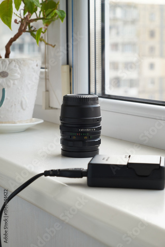 Camera lens and battery charger on a windowsill, natural light, photography gear setup at home with minimal composition and soft background
