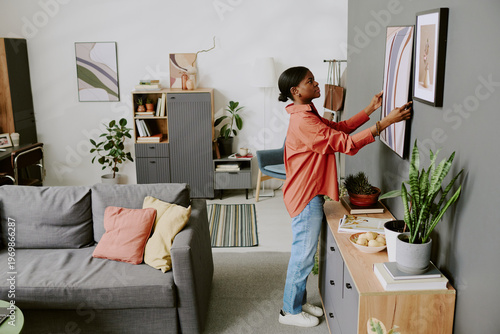 Photography Young Black woman arranging framed artwork on wall in living room, styling home interior for decor update and organization concept