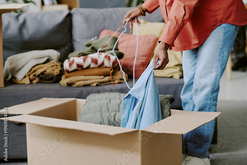 Tableau sur toile Black woman packing folded clothes into cardboard box while sorting hangers at home, preparing donation or relocation