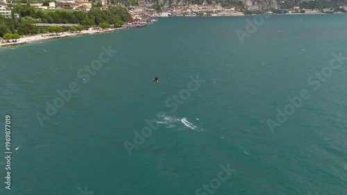 Beautiful 4K drone footage of a kite surfer cruising across Lake Garda, Italy, with stunning landscapes in the background. Filmed on a sunny summer day with clear blue skies, sparkling water, and pano