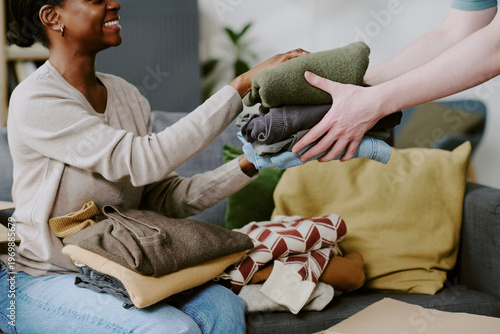 Photos African American young adult woman receiving folded clothes from helper while sorting donation items at home