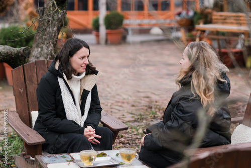 Two pretty caucasian women in their 40s wearing warm clothes spending time outdoors in autumn park cafe. Mature females enjoying personal time, talking, drinking tea, relaxing and breathing fresh air.