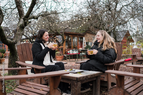 Two pretty caucasian women in their 40s wearing warm clothes spending time outdoors in autumn park cafe. Mature females enjoying personal time, talking, drinking tea, relaxing and breathing fresh air.