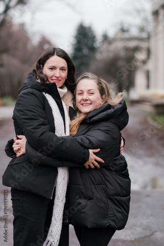 Two pretty caucasian women in their 40s wearing warm clothes walking outdoors in autumn park. Mature female best friends enjoying free leisure time, talking, hugging, relaxing and breathing fresh air.