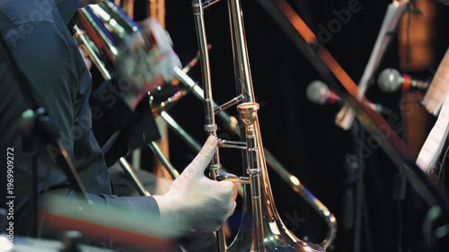 Close-up of a professional trombonist playing in an orchestra. The focus is on the instrument hands, with dynamic stage lighting creating reflections colorful bokeh on a dark background. Moody