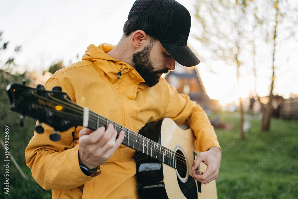 Naklejka premium Man plays guitar outside in yellow coat and black cap during golden hour