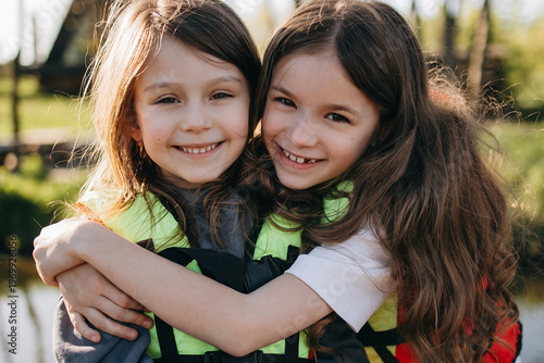 Two happy girls hug each other smiling brightly outdoors near water wearing life jackets