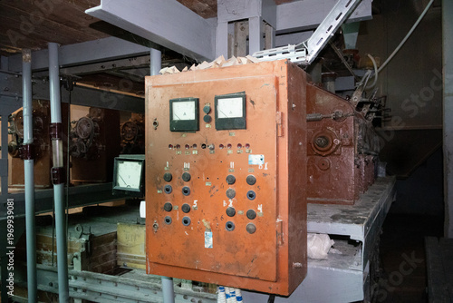 A weathered orange metal control box with analog dials and buttons mounted on large industrial equipment in a dark factory setting.