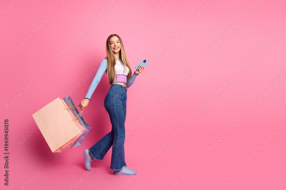Fototapeta premium Adorable young woman holding shopping bags and smartphone against pink backdrop radiating charm and happiness