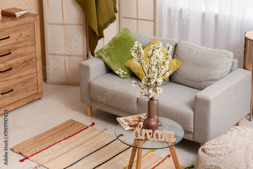 Vase of white blooming branches with word DREAM on coffee table near sofa in living room