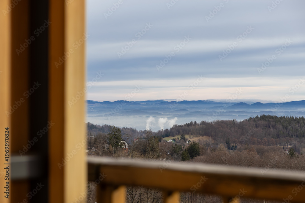 Fototapeta premium Serene panoramic misty mountain landscape featuring layered blue hills and bare forest trees, framed by a rustic wooden balcony, embodying peaceful nature scenery