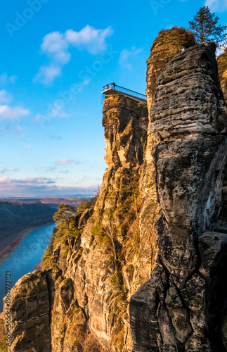 Lovely view of the Basteiaussicht perched on a sandstone rock needle of the Bastei formation, where visitors admire the Elbe Valley in golden winter sunrise light under a clear blue sky.