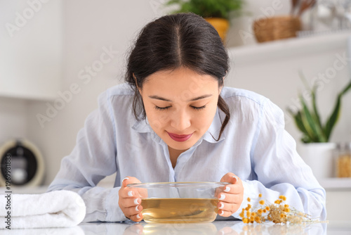 фотографія Young Asian woman doing steam inhalation at table in kitchen