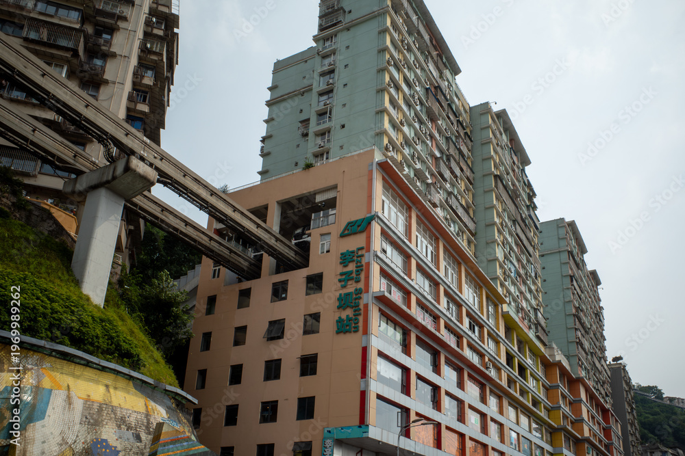 Obraz premium Urban high-rise buildings with colorful facades. Overhead train tracks cross a steep hillside. Greenery grows alongside concrete structures. Weathered walls show signs of urban wear