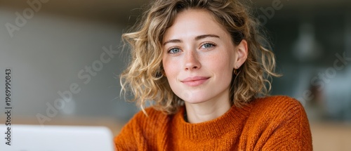 Young Woman With Curly Hair Wearing Orange Sweater Smiling At Camera Indoors Soft Background Light