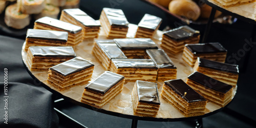 A close-up of a buffet display featuring layered pastries with chocolate glaze, canapes, and fruit arranged on a tiered stand.